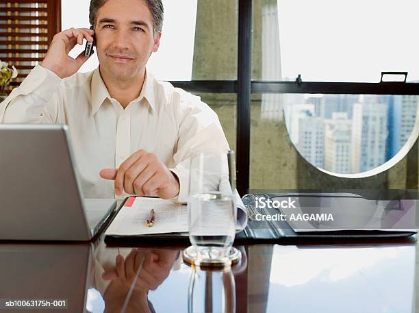 Businessman Sitting At Desk With Laptop Using Mobile Phone Stock Photo - Download Image Now