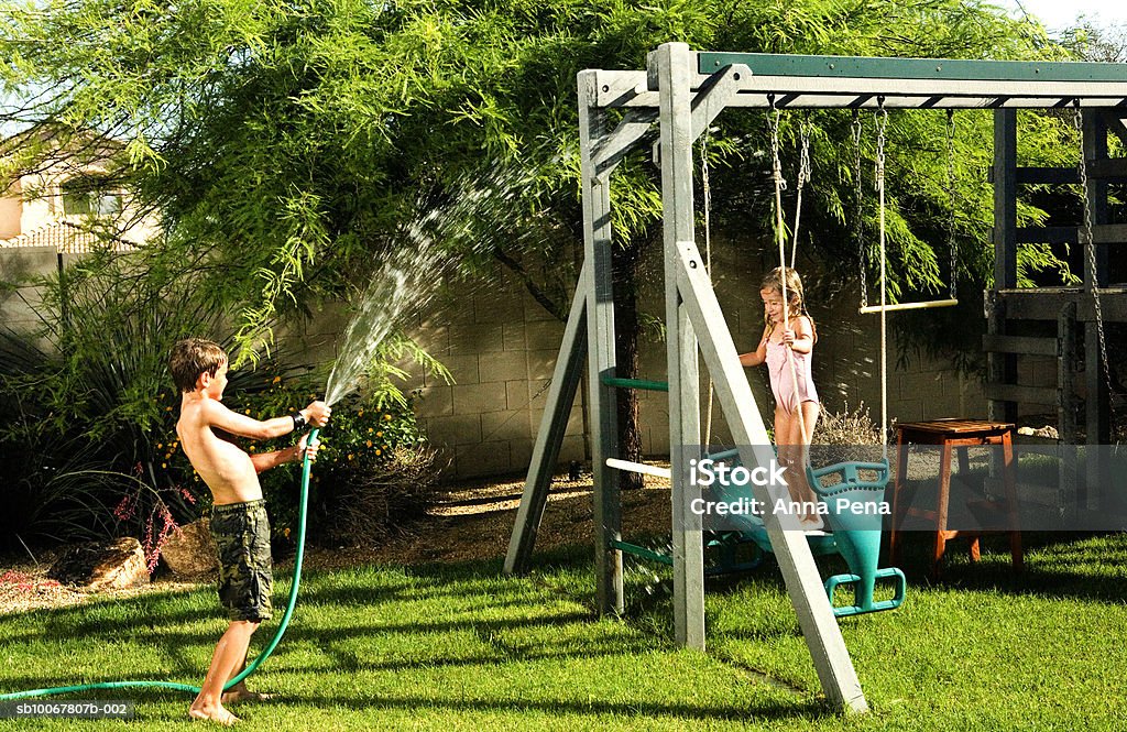 Boy (8-9) spraying water on girl (4-5) with hose - Foto stock royalty-free di Arizona