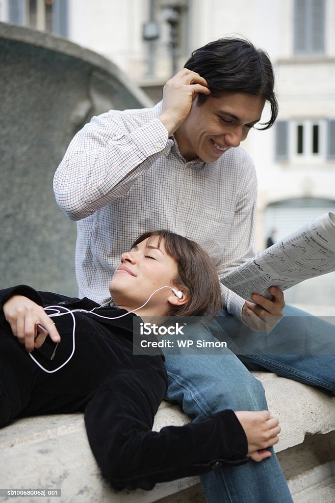 Woman listening to headphones lying head on man reading newspaper outdoors - Foto de stock de 20-24 Anos royalty-free