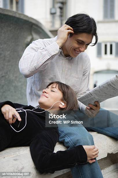 Foto de Woman Listening To Headphones Lying Head On Man Reading Newspaper Outdoors e mais fotos de stock de 20-24 Anos