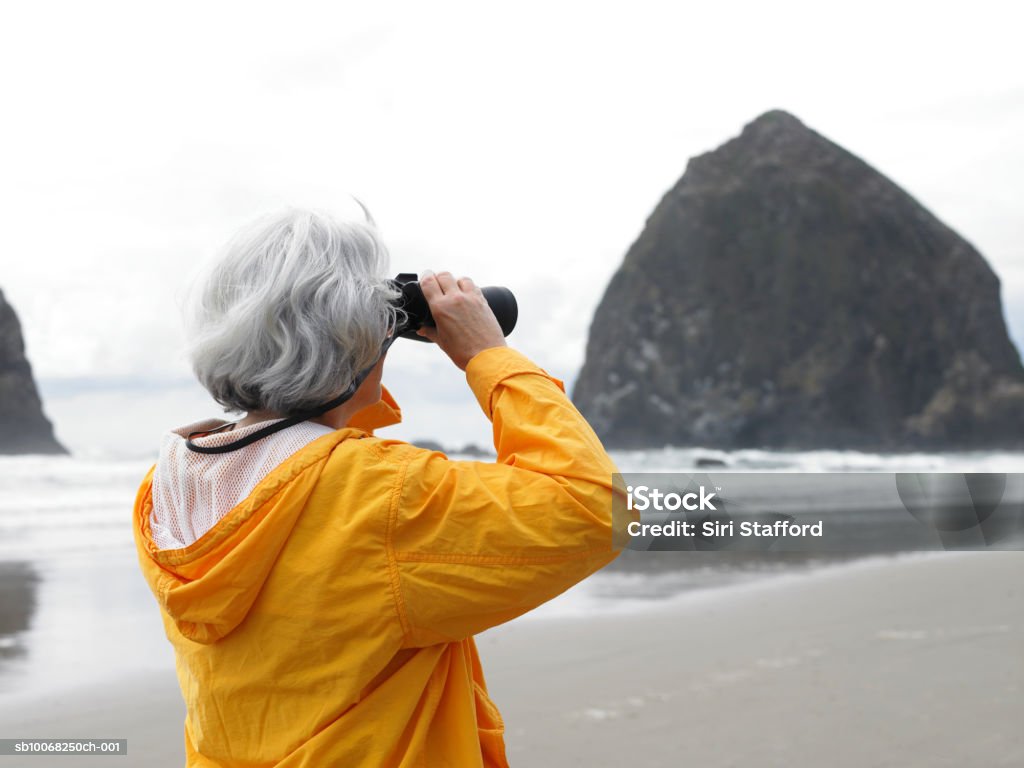 Senior woman looking through binoculars on beach Cannon Beach, Oregon, USA Binoculars Stock Photo