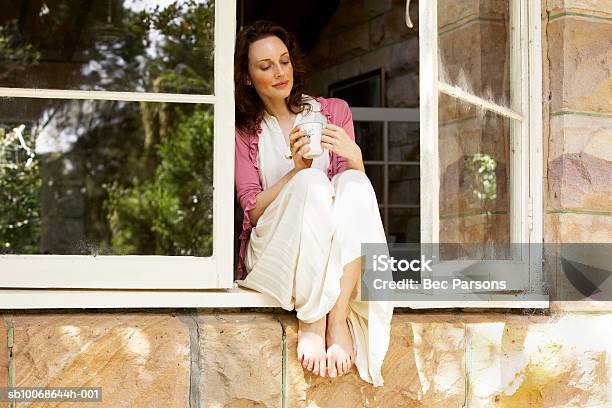 Mujer Sentada En La Ventana Con Una Taza De Café Foto de stock y más banco de imágenes de 30-34 años - 30-34 años, Adulto, Adulto de mediana edad