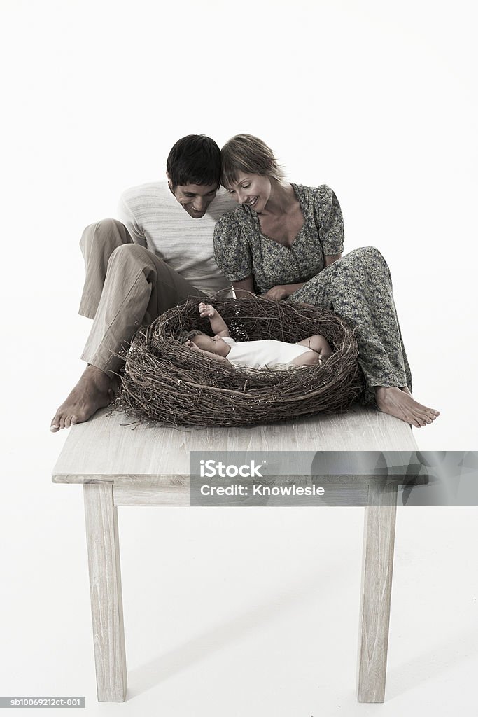 Couple looking at baby girl (2-5 months) lying in bird's nest on wooden table against white background  Baby - Human Age Stock Photo