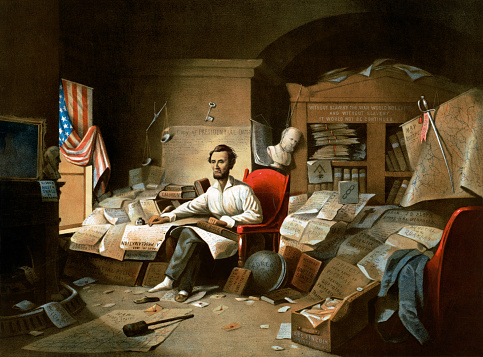 Vintage image depicts Abraham Lincoln writing the Emancipation Proclamation. In a cluttered study, Lincoln sits in his pajamas at work on the document, his hand resting on the Bible and the Constitution.