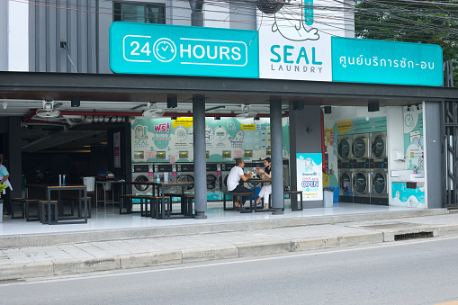 24h laundromat below condominium in Bangkok Chatuchak. View over street. Two young people are sitting at a table and are talking. Man is holding  an open book in hands and woman is holding a mobile in hand. In logo of laundrybar is a seal