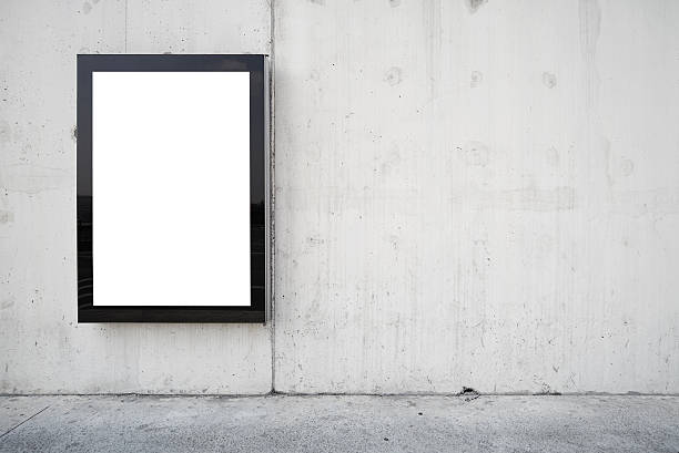 a blank billboard on a wall. Blank billboard on wall. Wall is made of concrete and gray coloured. Billboard is oriented vertically and standing on the left side of frame. Edges of billboard are black. Billboard is empty so you can write or add something on it. - Clipping path of billboard included. billboard posting stock pictures, royalty-free photos & images