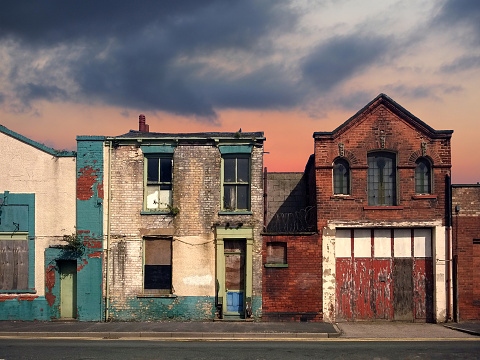 a deserted street of old abandoned ruined houses with bright peeling paint and crumbling brickwork in evening sunlight against a bright cloudy sunset sky redevelopment or fantasy concept