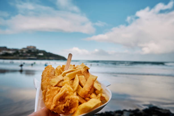 POV of a hand holding a carton of Fish and Chips at Towan Beach, Newquay, Cornwall on an Autumn day. stock photo