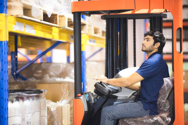 a man driving korklife in warehouse, worker job, work concept in store, loading cargo in factory, employee working, lifting goods. black hair guy stock photo
