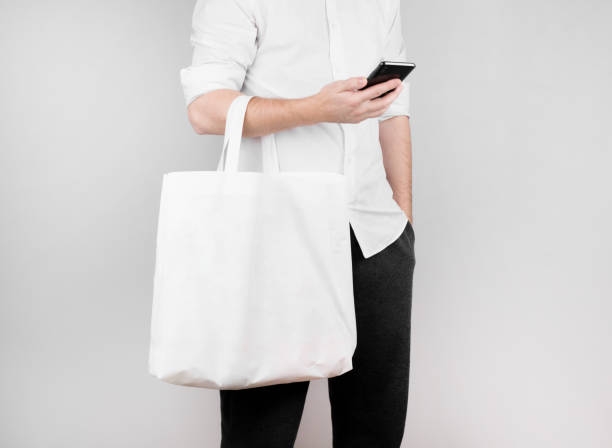 a man stands on a white background, reads the news on the phone and holds an ecological bag made of flax on his elbow. Ecology Concept stock photo