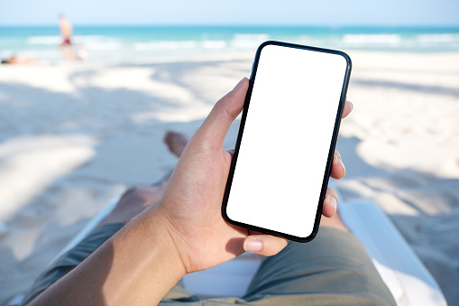 Mockup image of a man's hand holding white mobile phone with blank desktop screen while laying down on beach chair