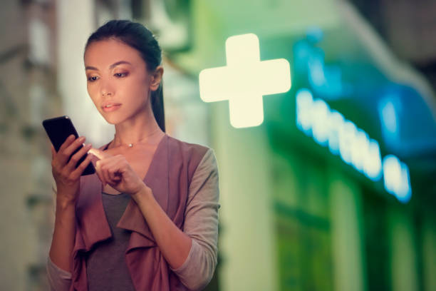 a young asian woman is locating a pharmacy store at night with her smart phone mobile device. communicate about open pharmacies during night time, using navigation to locate them, health, on call - sinal de emergência informação imagens e fotografias de stock