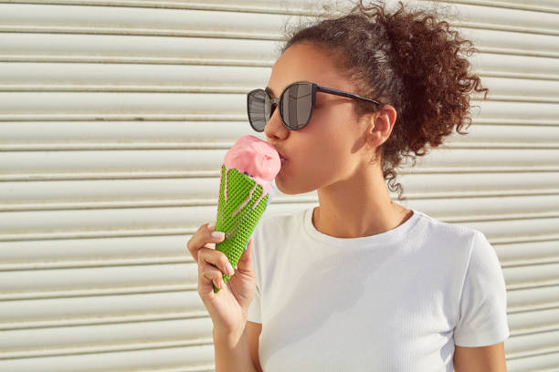 a young beautiful African-American girl in a white t-shirt and light jeans eats ice cream against a light wall on a Sunny day. stock photo