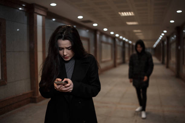 a young beautiful woman followed by a man in dark clothing with a hood on his head. a woman tries to dial the number of the rescue service on the phone. mosaics on the walls are not paintings stock photo