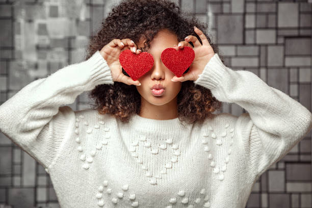 a young curly-haired African American girl in a white sweater holds two hearts in her hands . closes eyes with hearts and makes a kiss stock photo