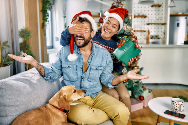 a young woman covering her boyfriend's eyes to surprise him and a dog sitting next to him stock photo
