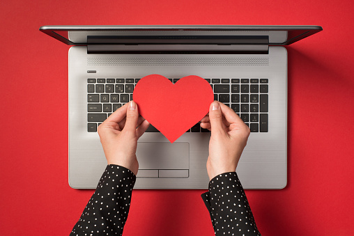 Above photo of grey laptop and hands holding a red paper heart isolated on the red backdrop