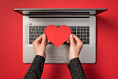 istock Above photo of grey laptop and hands holding a red paper heart isolated on the red backdrop 1337467349