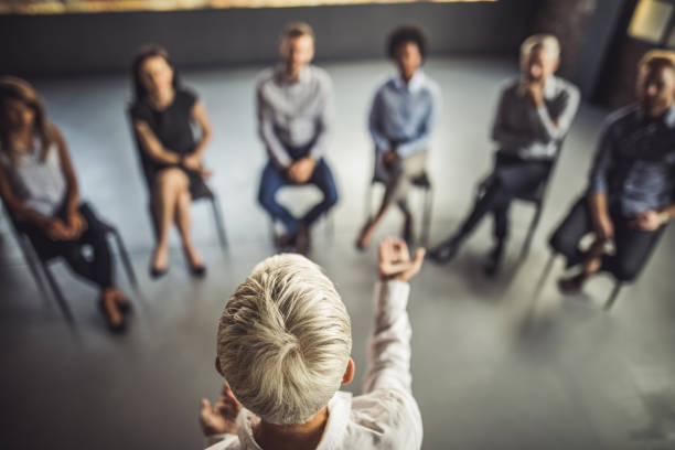 Above view of female instructor leading a business group therapy. High angle view of businesswoman talking to large group of her colleagues on a group therapy in the office. coach stock pictures, royalty-free photos & images