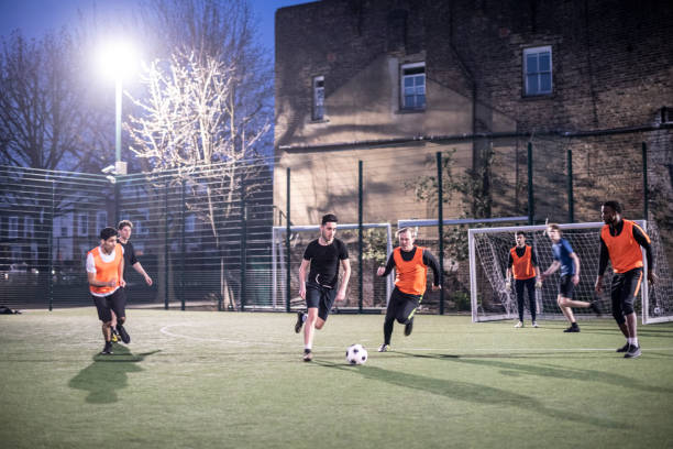 action shot of footballers on an urban pitch at night with two players running for the ball - soccer night imagens e fotografias de stock