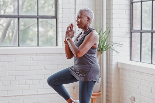 A retired black woman does yoga at home in her modern loft apartment to stay active and healthy. She is holding a standing balancing pose.