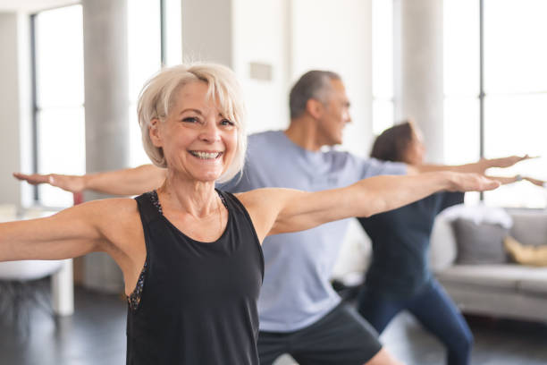 Active seniors in group fitness class A multi-ethnic group of seniors is attending a fitness class. They are indoors. The group is doing yoga. The individual in focus is a a beautiful and fit caucasian woman. She is smiling directly at the camera. Live Insurance stock pictures, royalty-free photos & images