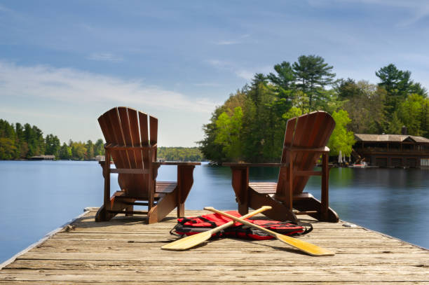 Adirondack chairs on a wooden dock facing ta calm lake Two Adirondack chairs on a wooden dock facing the blue water of a lake in Muskoka, Ontario Canada. Canoe paddles and life jackets are on the dock. A cottage nestled between green trees is visible. recreational pursuit stock pictures, royalty-free photos & images