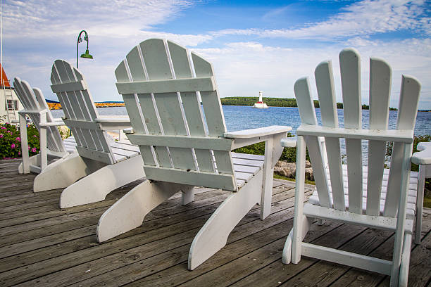 Adirondack Chairs On A Wooden Porch Overlooking A Lake Adirondack chairs line a wooden porch with the blue waters of Lake Huron and a lighthouse as the backdrop. Lifestyle shot that emphasizes the serenity of coastal living in Michigan's Upper Peninsula. This scene is on public grounds and not on private property. mackinac island stock pictures, royalty-free photos & images