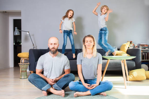 Adorable and lively siblings playing on sofa while parents sitting in lotus pose and meditate stock photo