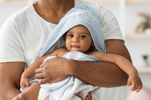 Adorable Little Black Infant Baby Wrapped In Blue Towel Relaxing In Father's Arms After Bath, Cute African American Toddler Child Looking At Camera, Enjoying Daddy's Care, Cropped Shot, Closeup
