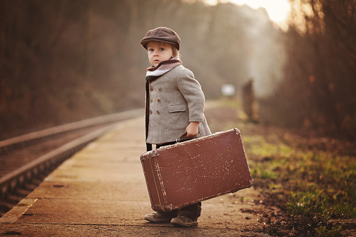 Adorable boy on a railway station, waiting for the train with suitcase and teddy bear, vintage look