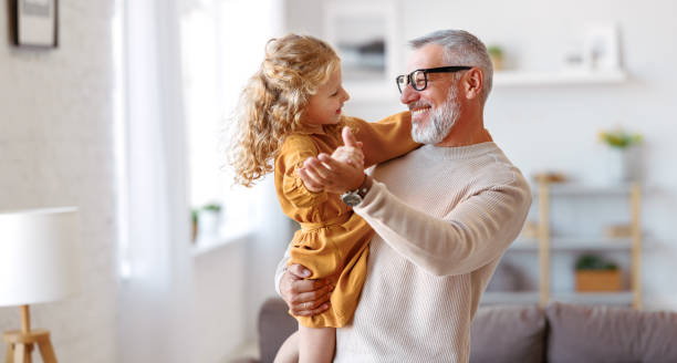 Adorable child girl and positive grandpa holding hands while dancing together in living room stock photo