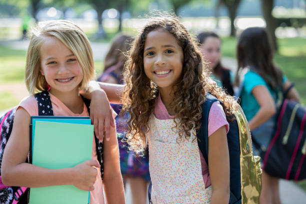 Adorable elementary age best friends wait outside school Caucasian and African American girls are smiling and looking at the camera while waiting outside school together. Students are wearing backpacks and holding homework assignments, dental braces photos stock pictures, royalty-free photos & images