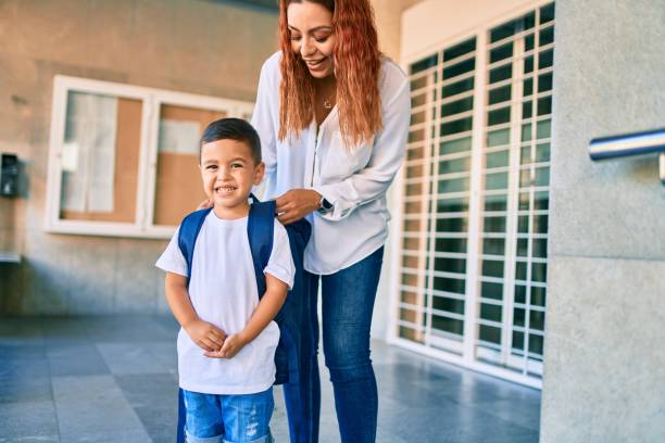 Adorable latin student boy and mom at school. Mother preparing kid putting up backpack. stock photo