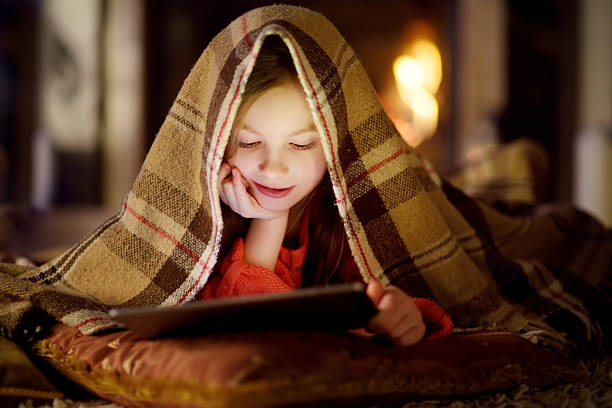 Adorable little girl using tablet by fireplace on Christmas evening Adorable little girl using a tablet pc by a fireplace on warm Christmas evening christmas story telling stock pictures, royalty-free photos & images