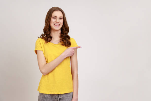 Adorable teenager girl with red curly hair wearing yellow T-shirt pointing to blank copy space and sincerely smiling, side empty wall for promotion. stock photo