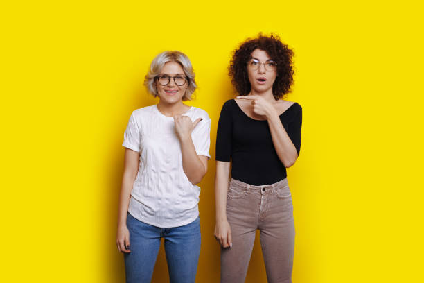 Adorable women with curly hair are smiling at camera and pointing at each other on a yellow studio wall stock photo