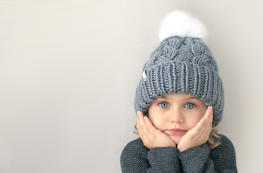 Adorible child girl with big blue eyes in grey wool knitted sweater and hat with white pompom looks at camera with hands on cheeks. Place for text, mock up, banner. Grey background.Handmade clothes