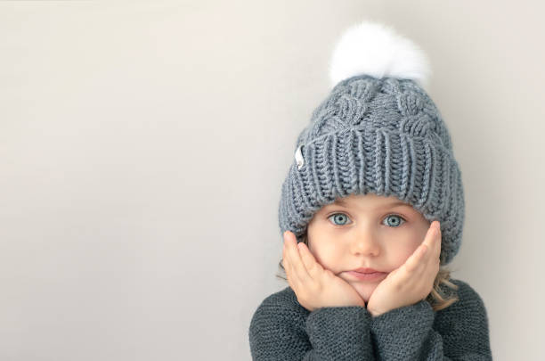 Adorible child girl with big blue eyes in grey wool knitted sweater and hat with white pompom looks at camera with hands on cheeks. Place for text, mock up, banner. Grey background.Handmade clothes Adorible child girl with big blue eyes in grey wool knitted sweater and hat with white pompom looks at camera with hands on cheeks. Place for text, mock up, banner. Grey background.Handmade clothes kids stock pictures, royalty-free photos & images