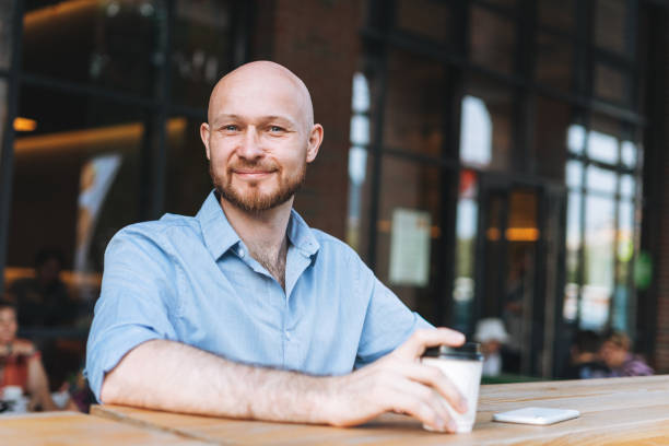 Adult bald smiling attractive man forty years with beard in blue shirt businessman using mobile phone with paper cup of coffee at cafe stock photo