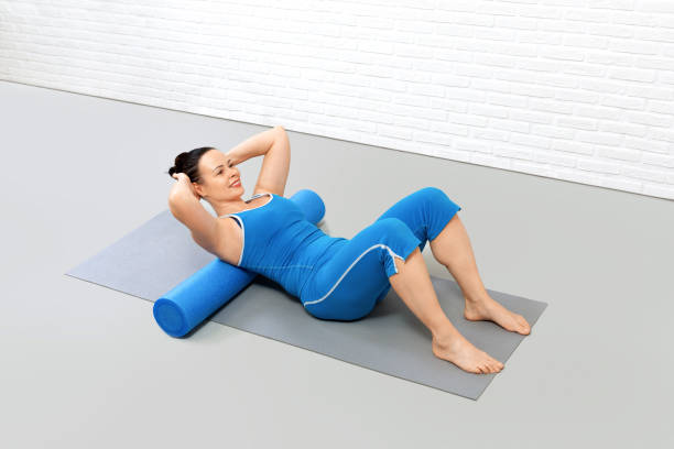 Adult caucasian woman in blue sportswear lies on a foam roller on the mat and stretching her upper back in fitness studio, top view. Pilates practice. Adult caucasian woman in blue sportswear lies on a foam roller on the mat and stretching her upper back in fitness studio, top view. Pilates practice. Workout indoors, good posture, thoracic spine, trainer, body extension, health, wellness concept. pelvic floor stock pictures, royalty-free photos & images
