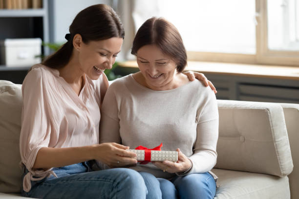 Adult daughter greeting mother with birthday at home Overjoyed grownup daughter sit on couch with smiling senior mother greeting giving wrapped gift, caring adult girl child congratulate make birthday surprise to middle-aged mom, celebrating at home birthday present stock pictures, royalty-free photos & images