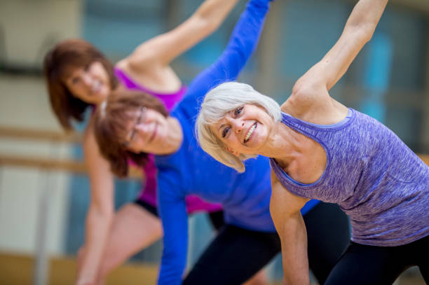 Adult Fitness Class Three Caucasian women are indoors in a health center. They are wearing casual athletic clothing. They are all smiling at the camera while stretching. 50 59 years stock pictures, royalty-free photos & images