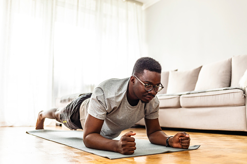 Confident muscled young man wearing sport wear and doing plank position while exercising on the floor in home interior.