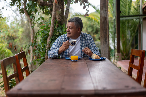 Adult Latin American man at a farm having breakfast at an outdoors dining room and drinking a cup of Colombian coffee