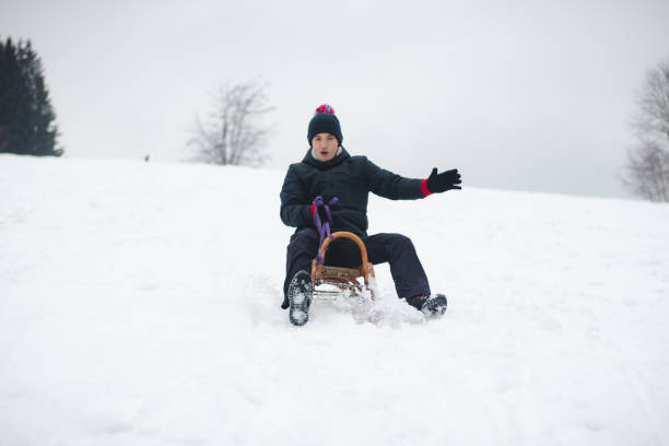 adult man in winter black clothes enjoys a ride on a wooden sledge with a smile and tries not to fall off it. Foot braking. Use of fresh powder for a fast ride. Winter season in the Czech Republic stock photo