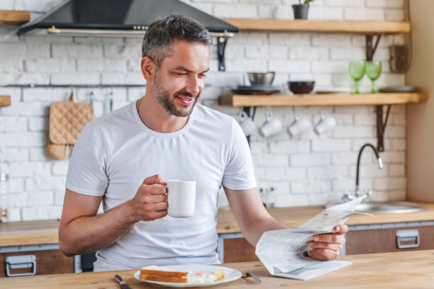 uomo adulto che legge il giornale e sorride mentre beve caffè e fa colazione in cucina - young man read newspaper foto e immagini stock