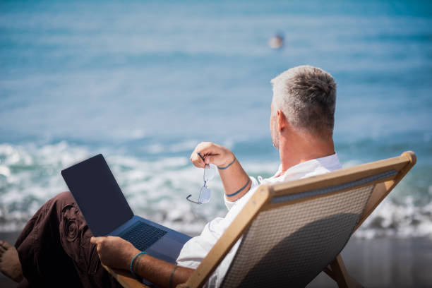 Adult mature man working on his laptop next to the beach Gray-haired man is sitting next to the blue sea in a lounge chair and working on his laptop nomadic people stock pictures, royalty-free photos & images