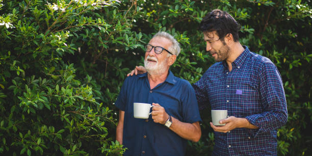 adult son and elderly senior father talking together and holding coffee cup in the garden, happy morning lifestyle of man generation family, love day at home stock photo