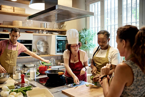 Adult Students Working with Chef in Cooking Class Senior woman in white toque and apron standing at kitchen workstation instructing students as they prepare vegetable ingredients. cooking class stock pictures, royalty-free photos & images